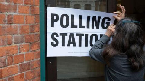 A woman sticks up a large sign reading "polling station" on the door of a church hall. 