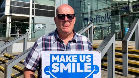 Man with very short hair wearing a grey, red and black shirt holding a sign saying "Make us Smile" in white lettering on a blue background. He is standing at the bottom of steps from the front door of the glass-fronted Suffolk County Council offices.