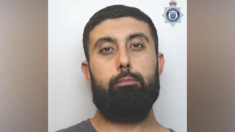 Asad Hussain, a man with short dark hair and a large black beard, poses in a police mugshot.