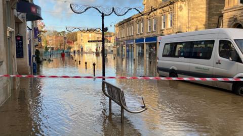 A town's high street submerged in water. There is red tape blocking access to the high street and a white Wiltshire Council van is driving through the flood water.