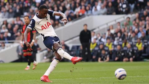 Pape Sarr in white and blue Spurs kit leans back slightly as he takes a right-footed shot, with the ball in shot skimming along the turf in front of him