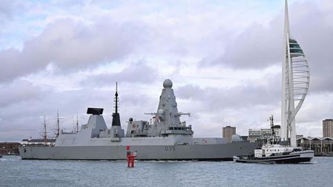 HMS Dragon, a large grey destroyer ship, sits on the water in Portsmouth Harbour, under a cloudy sky.