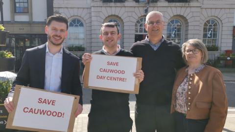 Four people standing outdoors in front of a building with arched windows, holding red‑and‑white signs reading “SAVE CAUWOOD!” and “SAVE CAUWOOD DAY CENTRE!”.