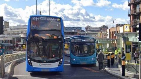 A double decker bus and a single decker bus on a road at traffic lights 