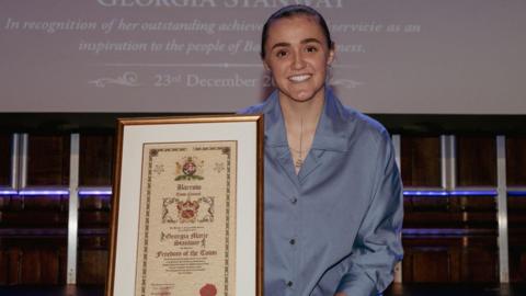 Women in a blue blouse with brown hair wearing a necklace. She is smiling and holding a certificate declaring her 'Freedom of the Town' award.