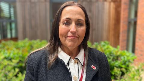 A woman with long dark hair smiles at the camera outside an officer building. She is wearing a dark grey jacket with a union jack lapel badge, and a red lanyard over her white blouse. The blurred background includes green foliage from a bed of shrubs.