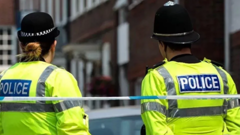Two police officers in uniform stand with their backs to the camera behind police tape. They are stood in a street with buildings visible behind them.