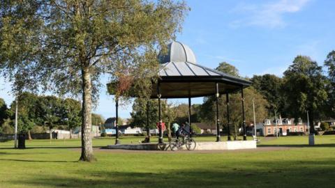 A bandstand in green parkland. There are a few trees and some children playing on their bikes.