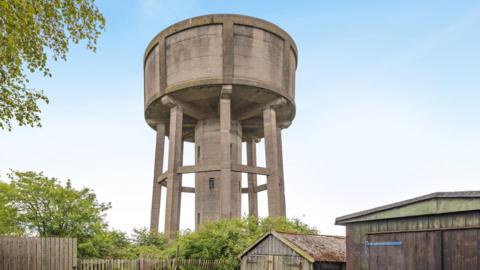 The water tower which is a round, concrete structure towering above the treeline and some wooden sheds. It has a central column down the centre with windows and about seven slimmer legs around it. It has a large, round top which is windowless. The sky above is blue and cloudless.