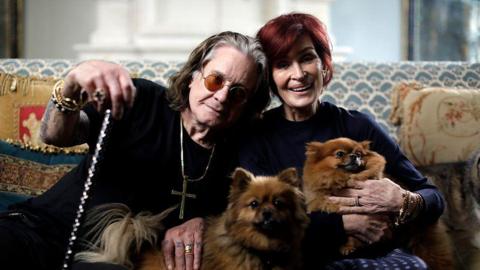 A photo of Ozzy and Sharon Osbourne sat on a sofa together. Ozzy is sat on the left and leans in towards Sharon so their heads are touching. He has shoulder-length grey hair and wears a black shirt and a large silver cross on a chain around his neck. He is holding onto a silver cane. Sharon has cropped red hair and wears a black top. There are two small dogs on their laps, one of which Sharon holds in her arms.