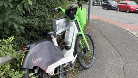 A battered looking lime bike leaning against a street sign on the side of a road.