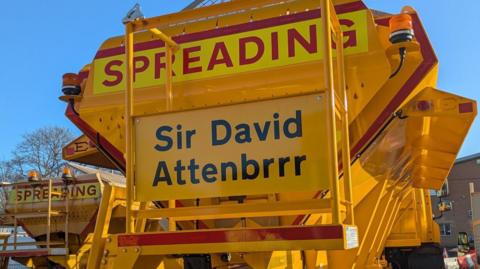 Yellow gritting vehicle with Sir David Attenbrrr on its rear.