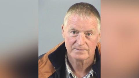 A elderly man wearing a brown jacket and checked black and white shirt.