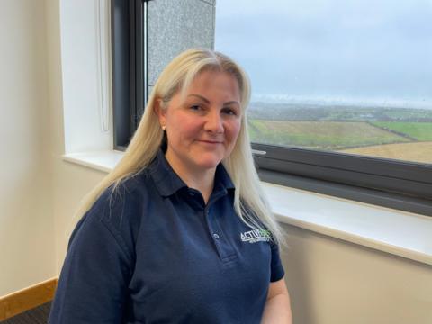 Joanne Whyatt wearing a blue navy polo shirt smiling at the camera whilst sitting by a window.