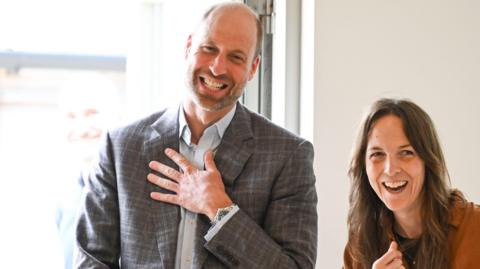 Managing Director Rachel Phillips and the Prince of Wales talk indoors near a bright window with one hand on his chest, both facing toward the camera.
