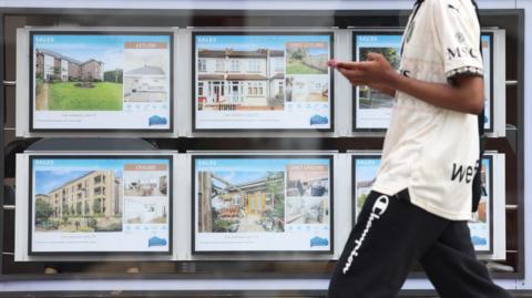 A man walks past an estate agent window filled with six bright digital screens displaying homes. 