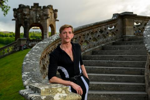 Stephen Libby, a man with blonde hair and a well groomed moustache standing on a grand outdoor staircase, which he is leaning on with his left arm. He is wearing a striking black jumpsuit with a deep v-neck and white binding down the sides.