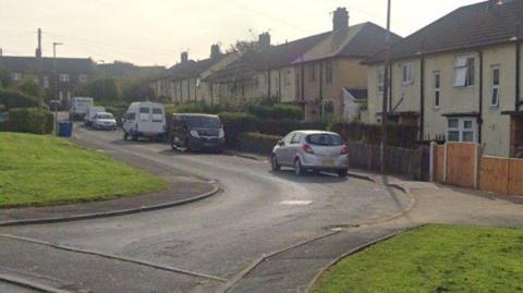 Google Earth photograph of Pilmuir Road in Blackburn. Cars and vans are parked along the road, with a row of semi-detached houses and more homes at the top of the road. Two grass verges can be seen on either side of the entrance to the road.