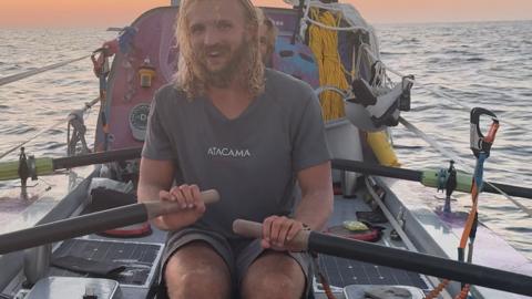 Man with long hair sits in a rowing boat, behind him is equipment, dusk is falling over the Pacific Ocean