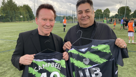 Campbell and Hassan stand together holding the shirts which are on hangers. Both are wearing black tops and jackets and are stood on the football pitch.