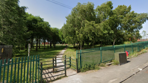 A playing field. In the foreground is a pavement and a length of green metal fencing with an entryway to the field. Behind the fencing there is a path, with trees either side of it