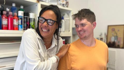 Simone and Alex are laughing together. Simone has long dark hair tied up and glasses, and is wearing a white shirt, and Alex has short brown hair and is wearing a yellow T-shirt.