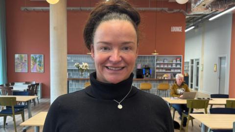 Laura is smiling at the camera in a cafe area, with tables visible behind her. She is wearing a black polo-neck jumper, a silver necklace, and has dark hair which is tied back in a bun. 