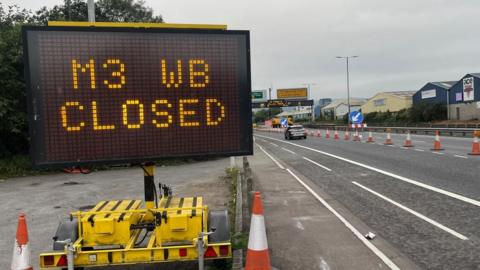 A dual carriageway has orange and white cones running down the middle dividing line in the road. There is a large automated sign in the foreground that says 'M3 WB CLOSED'.