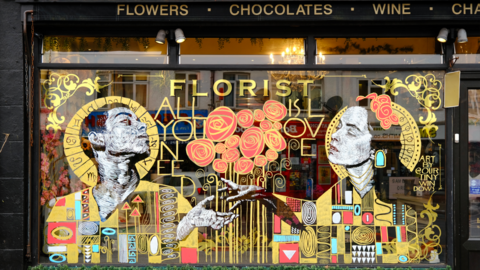 A lavish window display in gold and red coloured paint takes up the entirety of a shop window. It shows two figures who are depicted as religious-looking icons reaching out their hands to each other with red flowers between them.