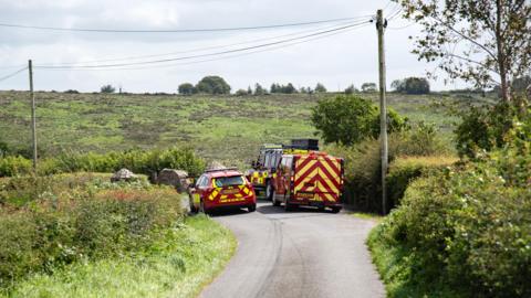 Red and yellow striped emergency vehicles are parked on a country lane which is surrounded by green fields.  