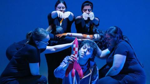 Six actors performing a scene of The Curious Incident of the Dog in the Night-Time. A male actor in the middle, wearing a lighter coloured track suit is holding small rocket, five other actors, all dressed in black, have their arms out and they are holding teach other. 