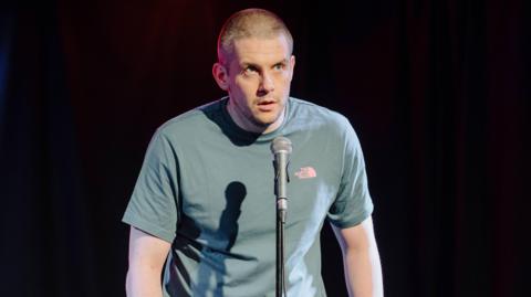 A closeup of a man with a buzz cut standing in front of a microphone, wearing a pale green North Face t-shirt. He looks out at the crowd, backed by total darkness. He looks apprehensive, likely about to drop the punchline of a joke.