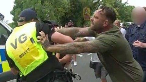 A tattooed man wearing a green T-shirt appearing to shove a police officer wearing a hi-vis top amid the backdrop of a protest.
