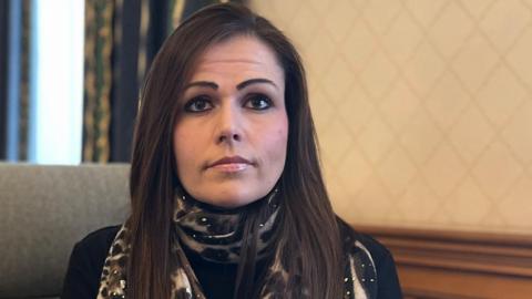 Lucy Domaille - a woman with long brown hair, looking at the camera. She is wearing a leopard print scarf with a black jumper underneath. 