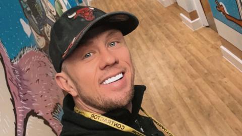 Adam Salisbury wearing a black baseball cap and black fleece standing in a hospital corridor with his artwork on the walls. He is smiling. 