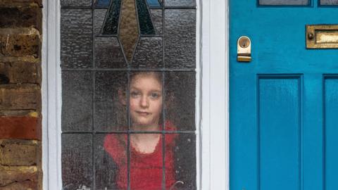 A little girl in a red t-shirt looks through a textured and stained glass window next to a blue door and a red brick wall. 