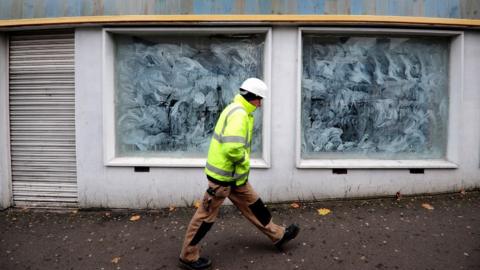 A man in a high-vis jacket, white hard hat and brown trousers walks past a close shop front, with shutters down on the door and windows covered in white paint. 