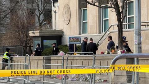 An image showing law enforcement members outside the US consulate building in Toronto, surrounded by caution tape. There is also a small memorial visible for the US soldiers killed in the Middle East during the ongoing war in Iran, with flowers laid around a sign with their photos.