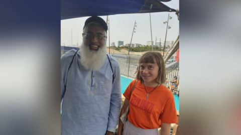 Mr Hussain is wearing a blue shirt and has a back pack on. He has a long, white beard and glasses and a cap. He is stood next to Greta Thunberg who is on his right as you look at the picture. She is shorter than him with an orange top on and a bag over her shoulder. She has a white skirt on.