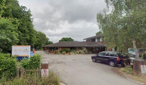 A Google image of the outside of a building, taken from the road. 5mph signs are on a low wall at the entrance and an Acorns sign on the left. The building is one and two stories high and surrounded by trees and a carpark.