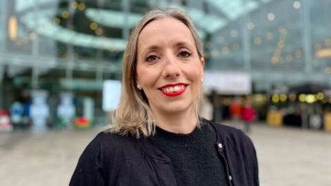 Sarah Taylor in a black top and black cardigan. She is smiling towards the camera and has blonde hair.