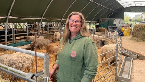 A farmer wearing a green hoodie smiling at the camera while she stands inside a metal barn with sheep behind her