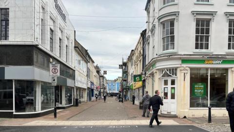 A view looking down King Street in Whitehaven. There is a Subway shop on the right with an empty unit on the left. Several signs look tired and dirty, with scaffolding visible further down the pedestrian street.