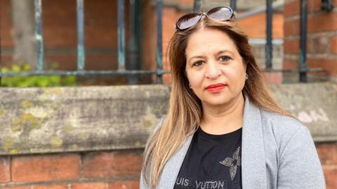 Rani Rawji stands outside the library. She is wearing a black top and grey jacket and has long hair and red lipstick.