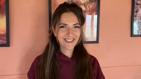 A woman with long dark hair smiles at the camera. She is wearing a burgundy hoody type top and is standing in front of a pale pink wall with framed pictures on.