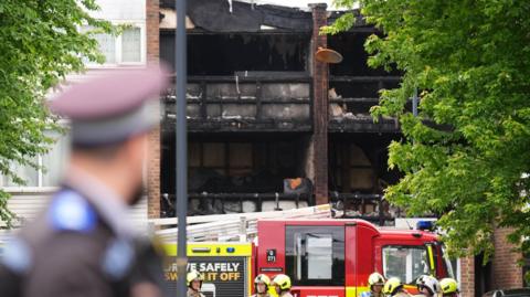 The scene in Tillett Close, Stonebridge, in Brent, north-west London, where a 15-year-old girl, two boys aged eight and four, and a 43-year-old woman died after a house fire in the early hours of Saturday. In the image you can see a police officer in uniform in the foreground, slightly out of focus. In the background there is a fire engine and  burned out flats,