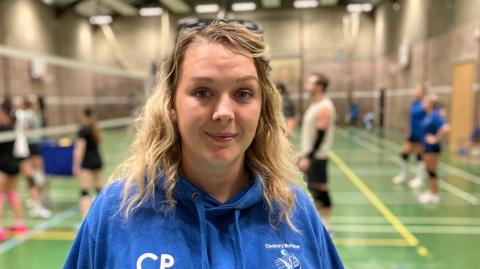 A blonde-haired woman with sunglasses on top of her head, wearing a blue hoodie in front of a green-floored sports hall with adults playing volleyball.