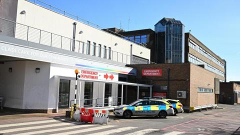 The front of a hospital building on a sunny day. A couple of police cars are parked outside. 