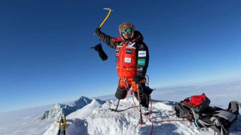 A man with prosthetic legs stands on the snowy summit of Mount Vinson in Antarctica. Below him is a sea of white cloud.