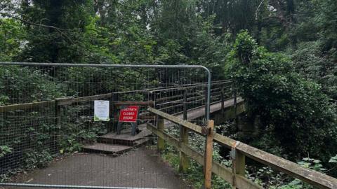 A metal gate is blocking the entrance of the wooden bridge at Cherry Tree Walk in Jesmond Dene. A red sign reading FOOTPATH CLOSED AHEAD is place behind the gate, where the narrow wooden footbridge begins. The structure is surrounded by trees and shrubs.